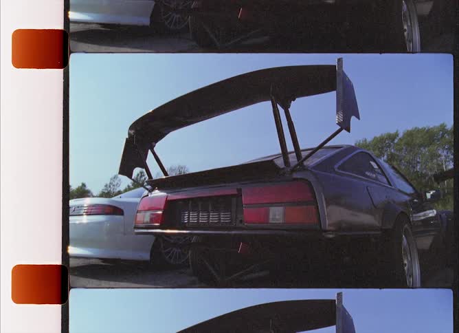 A sports car with an extremely large custom spoiler is parked outdoors next to another car