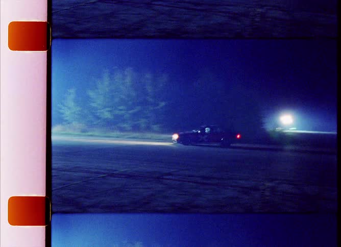 A vehicle with headlights driving on a track with trees and lights in the background