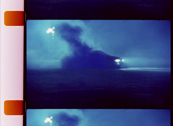 Smoke rises from parked car in foggy field under light towers