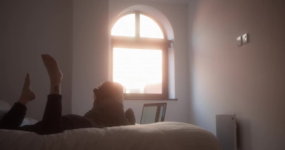 Woman lying on stomach on bed kicks her feet in air while she views laptop screen