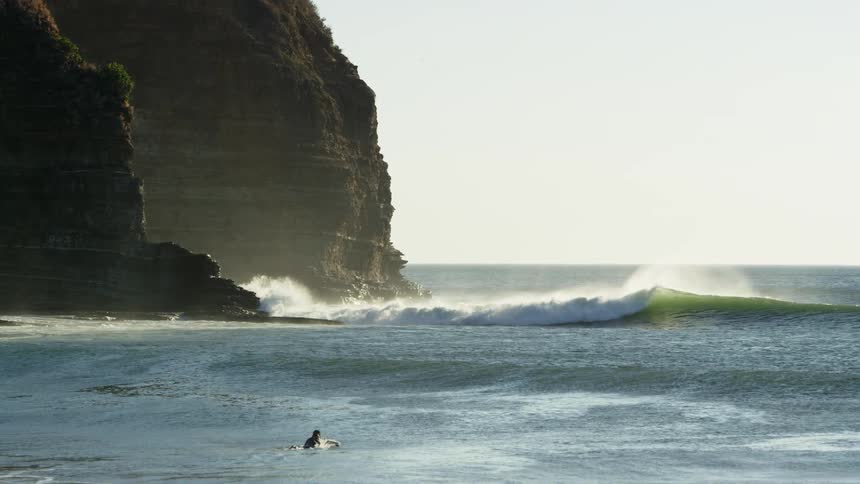 Surfer wearing a black wet suit swimming over a surfboard in the ocean near a cliff