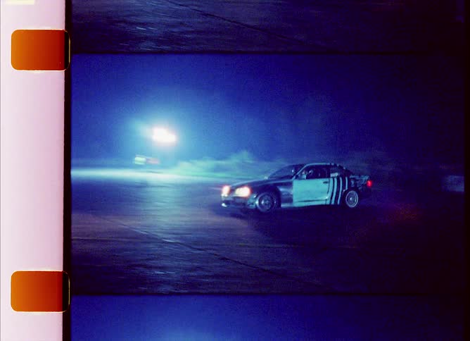 A vehicle with headlights driving on a track with spotlights in the background