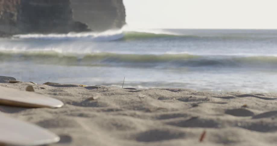 Two surfboards lay atop the sand on a beach as waves crash into the shore nearby
