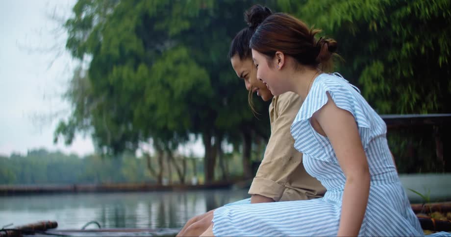 Couple sit on dock soaking feet in lake laughing talking and gesturing