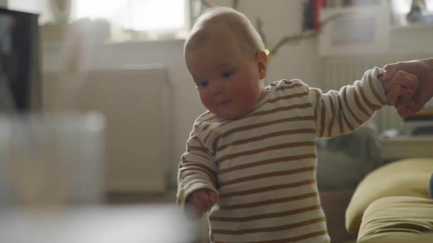 Mother sitting in a couch on the living room holds a baby hand to help him stand