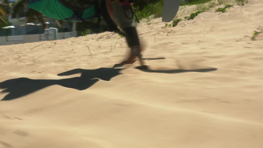 Man with a prosthetic leg walking on a beach carrying kitesurfing equipment