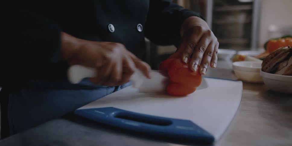 A female chef uses a large knife to slice a red bell pepper in half in a professional kitchen