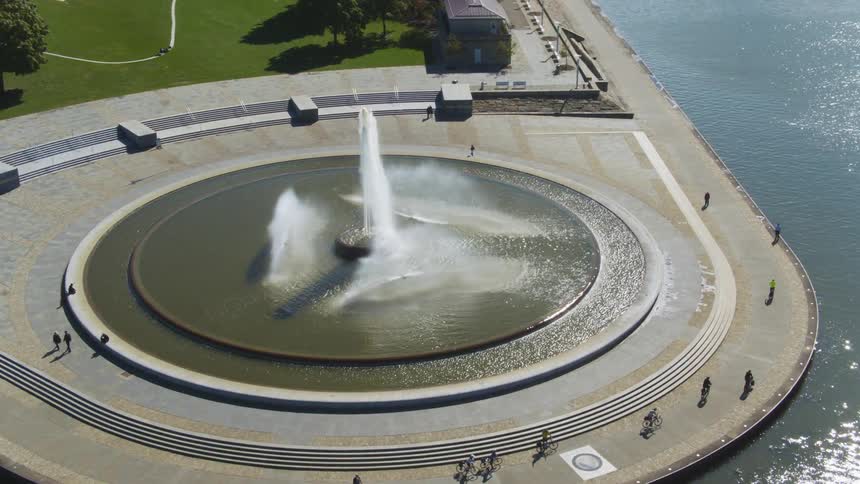 Pedestrians and bicyclists move along large fountain bordering water reflecting sunshine