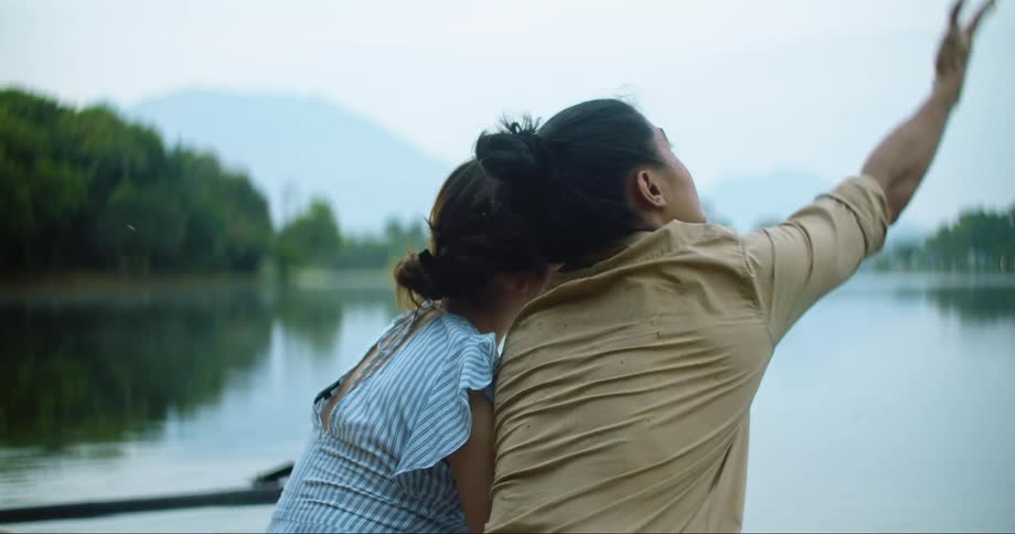 Couple leans against each other near lake reflecting trees on shore as man splashes water into air, mountains on the horizon