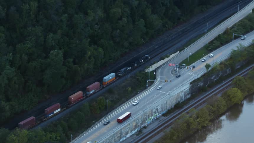 A cargo train moving on a track next to a road with vehicles and a river