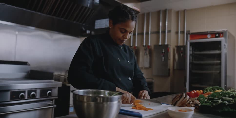A female chef uses a large knife to carefully cut vegetables as she prepares food