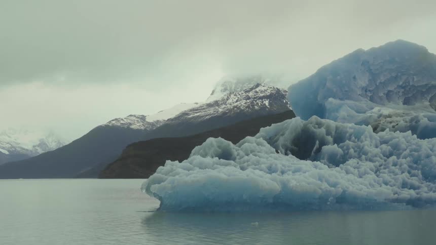 Iceberg floats in water with promontory backed by mountains in background under overcast sky