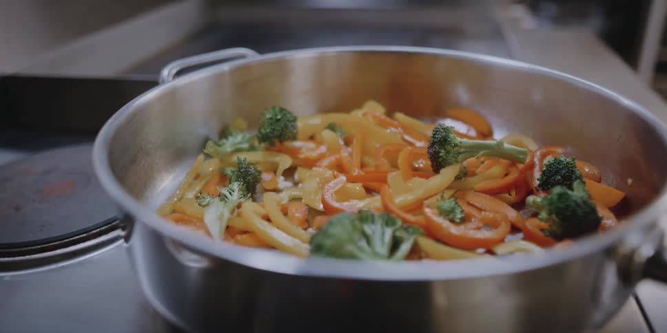 A woman stirs a pan of vegetables on the stove of a restaurant kitchen