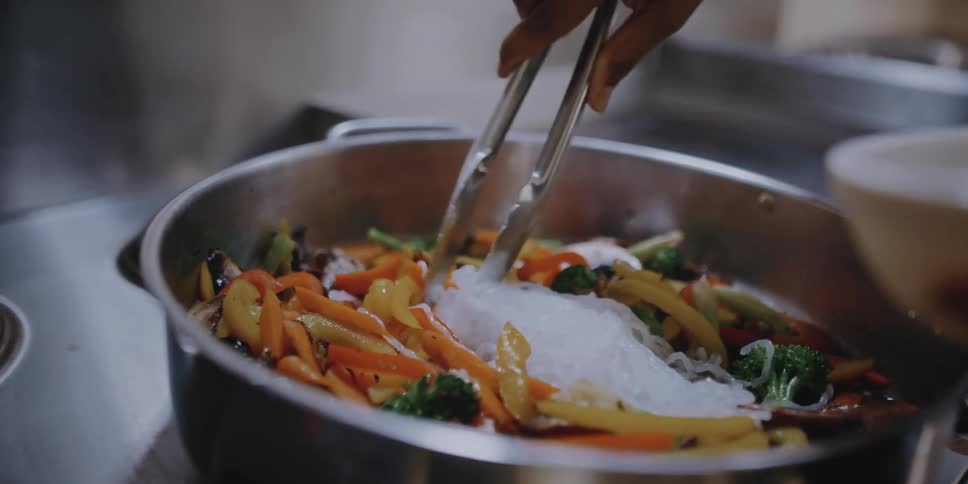 A restaurant chef uses tongs to take food from a bowl and place it in a large pan on the stove