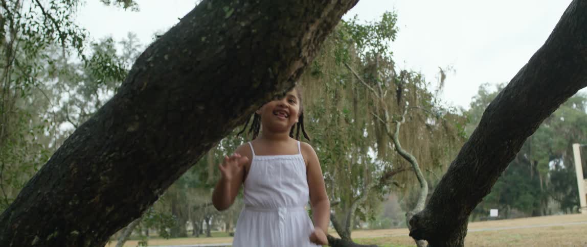A girl in a white dress runs laughing around a tree