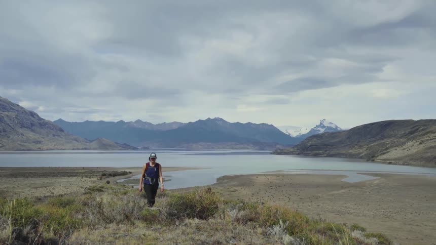 A person with a hat walking in a landscape with a mountain range the sky and clouds in the background