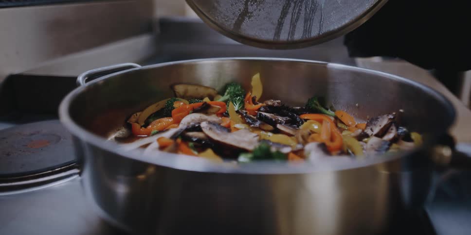 A chef lifts the lid on a pan in which she is cooking vegetables with mushrooms
