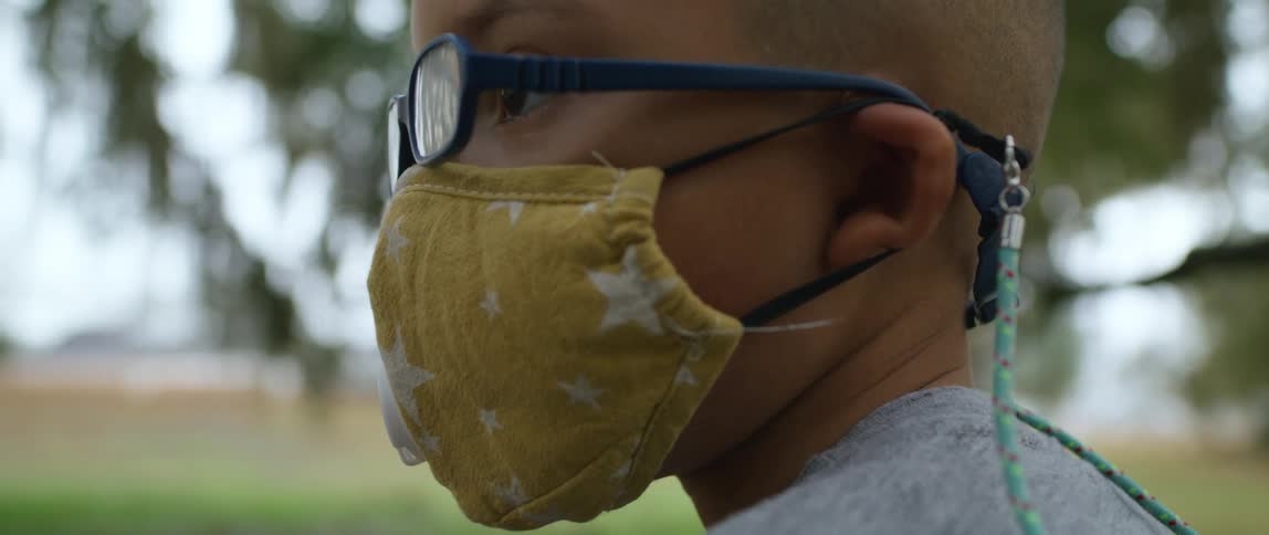 A young boy wearing a colorful face mask stands outdoors at a park