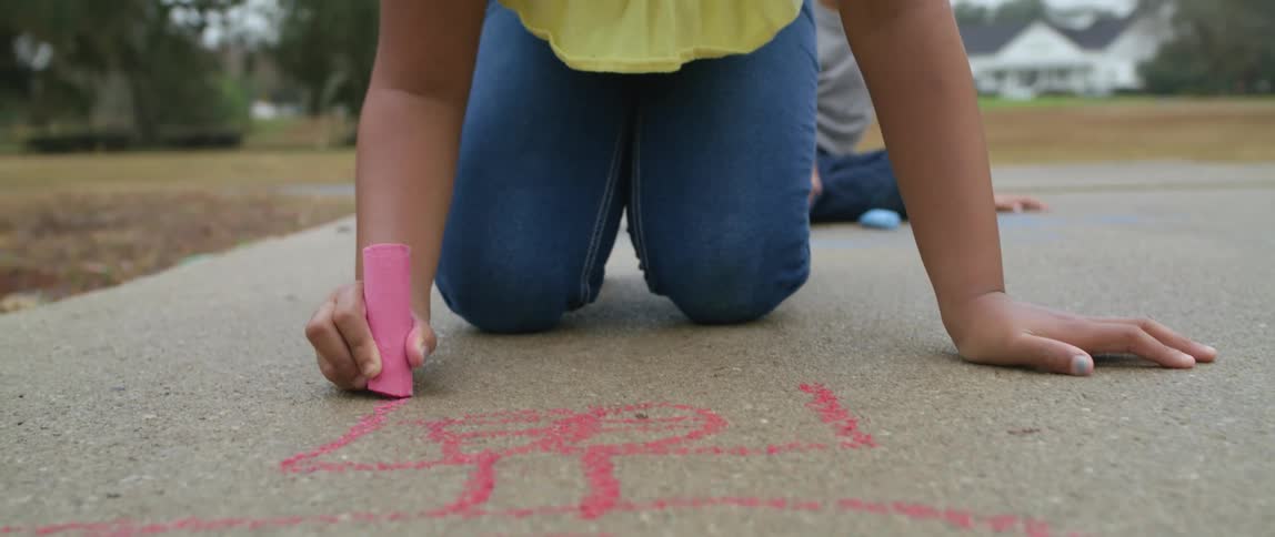 A young girl kneels on the sidewalk coloring it with chalk
