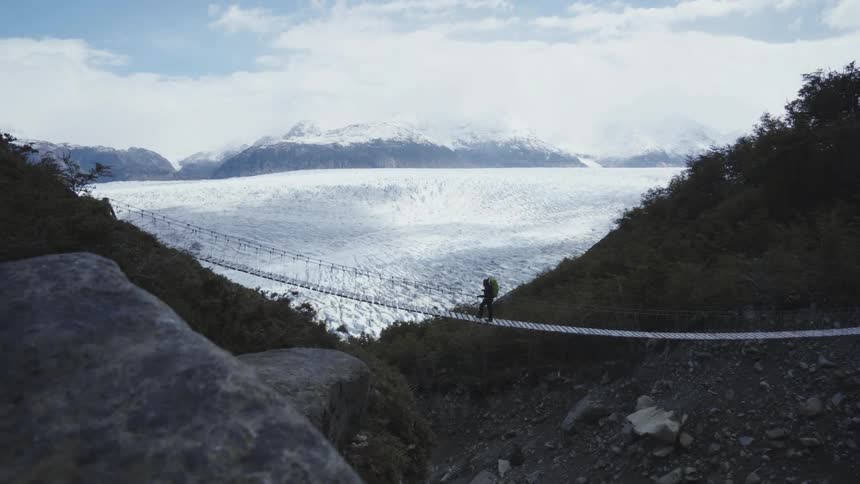 A person is walking across a swinging bridge with snow covered mountains in the distance