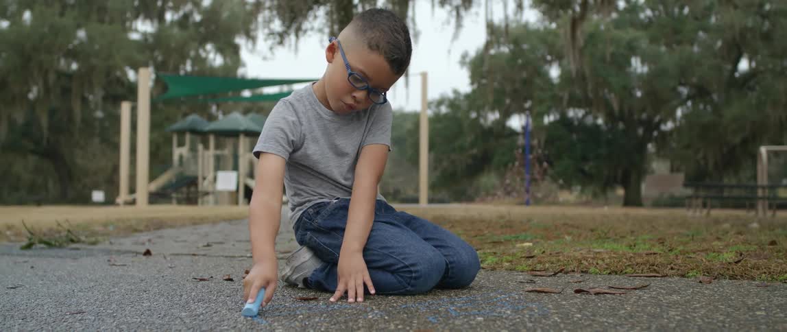 A young boy kneels on a walkway at a park while drawing on the ground with chalk