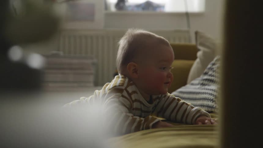 A baby in a room standing by a couch with a wall and window in the background
