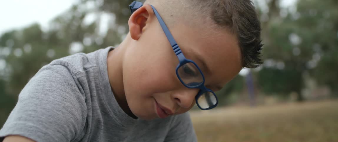 A boy with glasses drawing with chalk on the ground with the sky and clouds in the background