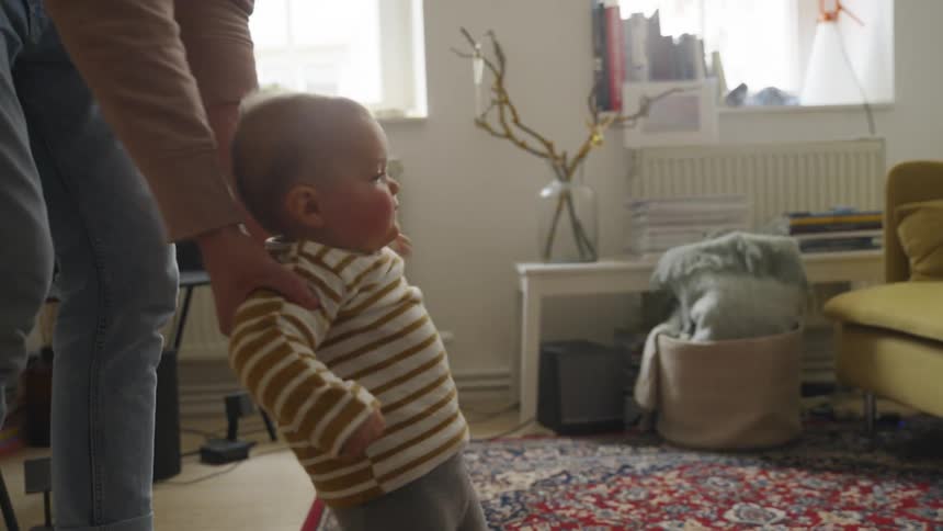 A woman helps a baby walk across a living room