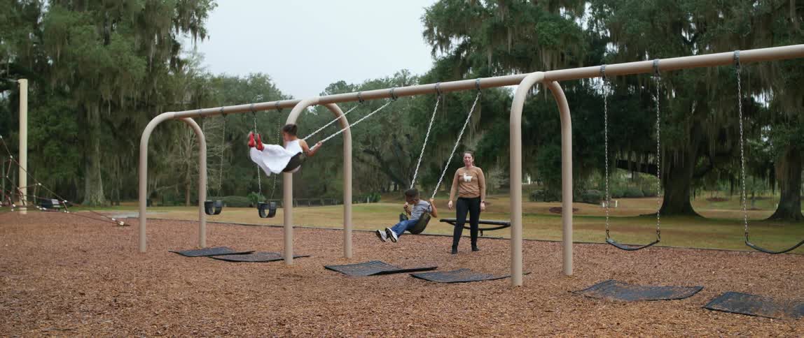 A woman pushing a girl on a swing with a boy swinging next to her with trees the sky and clouds in the background