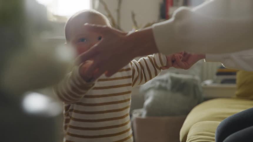 Blonde woman kneeling on furniture helps baby stand up by holding hands