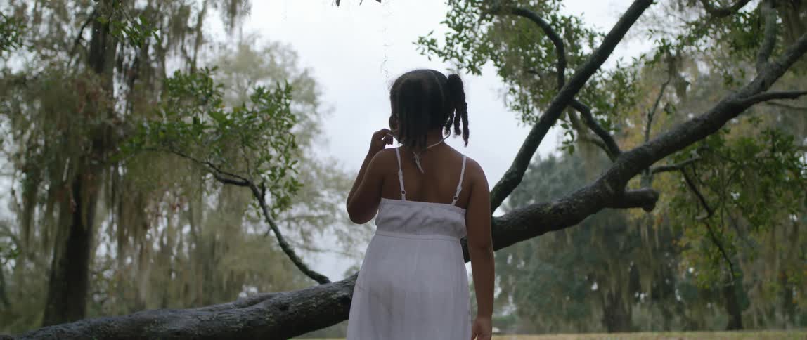 A young girl in a white dress turns and looks around at her surroundings in a grove of trees
