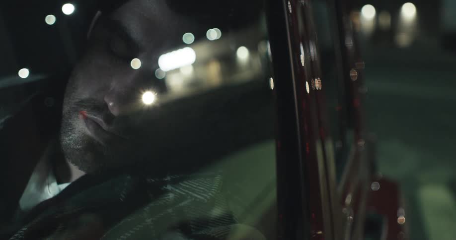 Man wearing plaid coat sleeps in car as street traffic and nearby building is reflected in glass