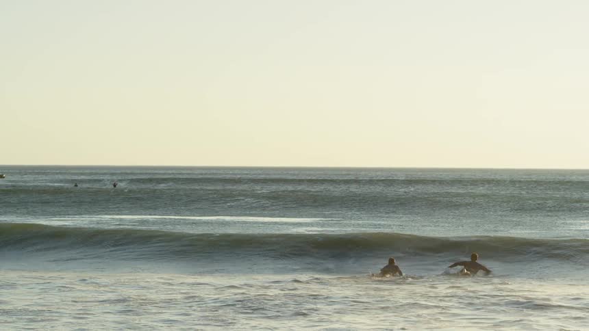 Two surfers swimming over a wave in the ocean next to a cliff at sunset