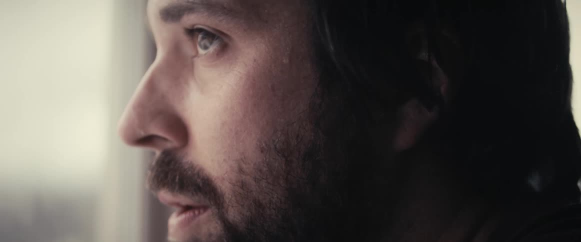 A bearded man looks around and wipes sweat from his face with the sleeve of his shirt