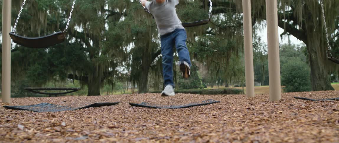 Boy jumps of swing in park near tree covered in moss