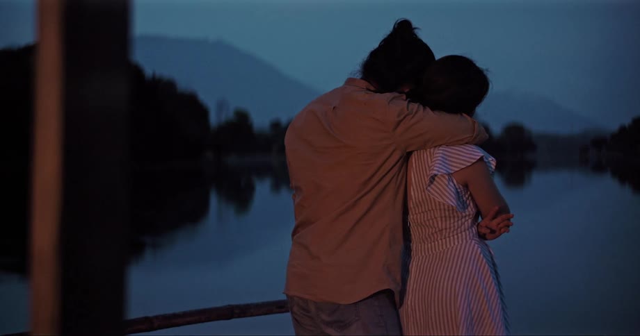 A man and a woman hugging with a pond trees the sky and clouds in the background