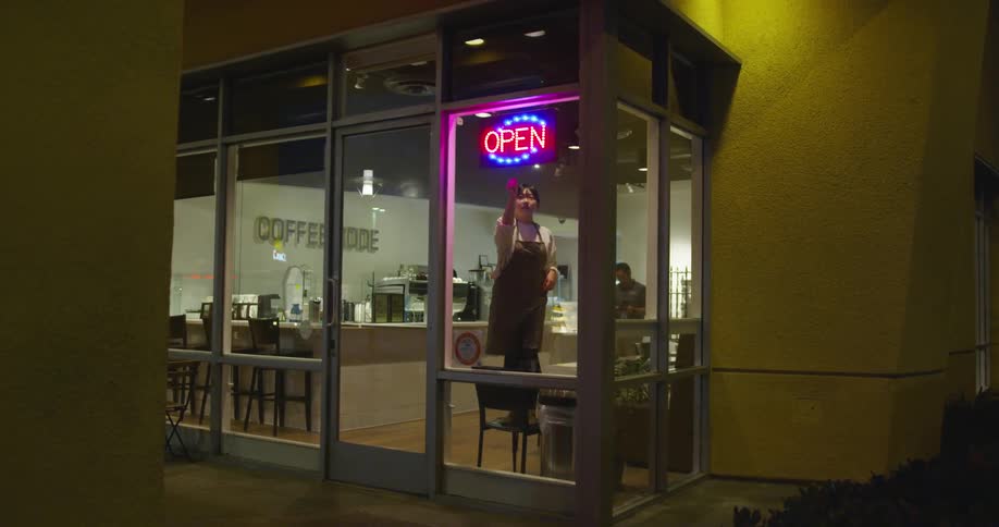 A young woman wearing an apron stands on a chair in order to turn on the open sign for a small coffee shop buisness