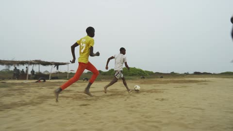 A group of barefoot boys play soccer in sandy soil on a cloudy day