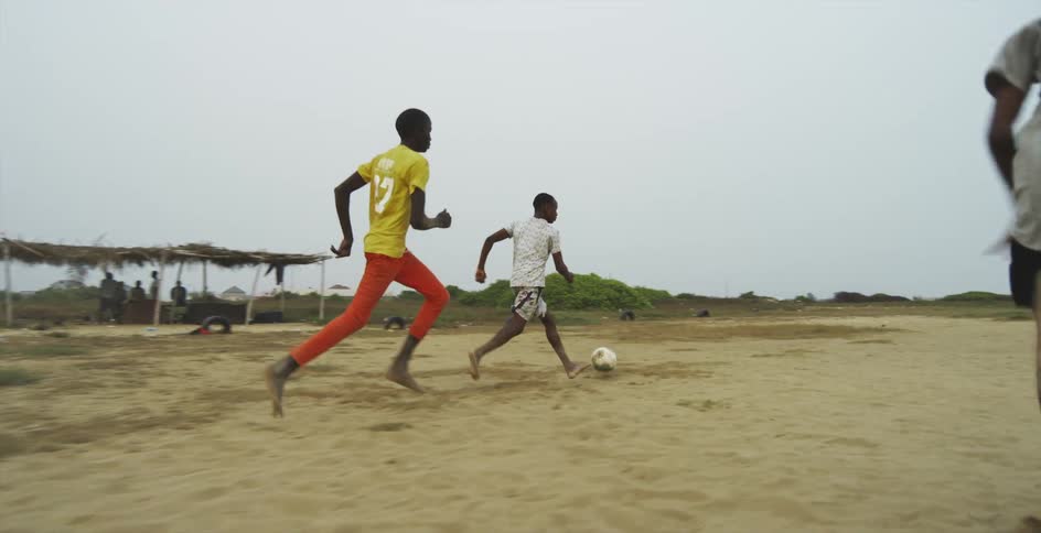 A group of barefoot boys play soccer in sandy soil on a cloudy day