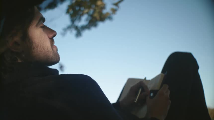 A man is sitting outdoors and writing on a journal