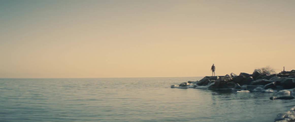 A man stands atop a rocky prominence along the shore of a large body of water