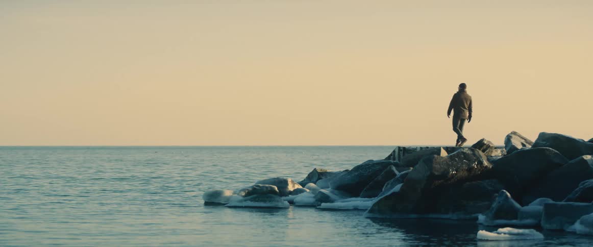 A man walks atop a rocky prominence at the shore of a large body of water