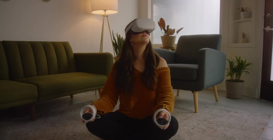 A woman wearing a virtual reality headset looks around while sitting cross legged on the floor of the living room of a house during the day