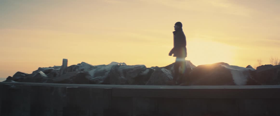 A man with a hat and jacket walking on a pier with the sunset the sky and clouds in the background