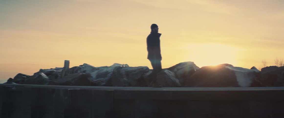 Man wearing jacket walks along causeway away from shore as sun on horizon turns sky red