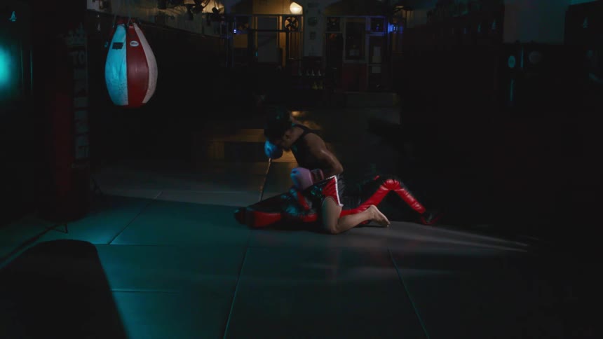 Fighter wearing boxing gloves kneels over cushion punching it in dimly lit gym near speed bag