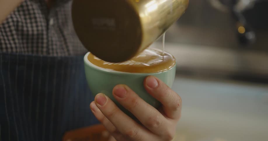 A baristas hand holding a cappuccino decorating the top with cream from a pitcher