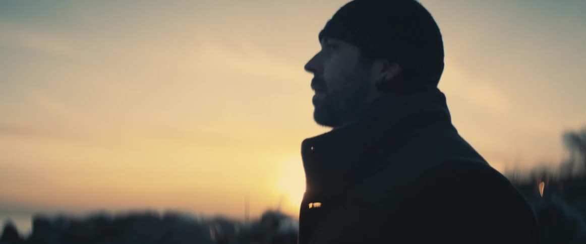 A man stands outdoors while looking off into the distance as the sun is low in the sky behind him