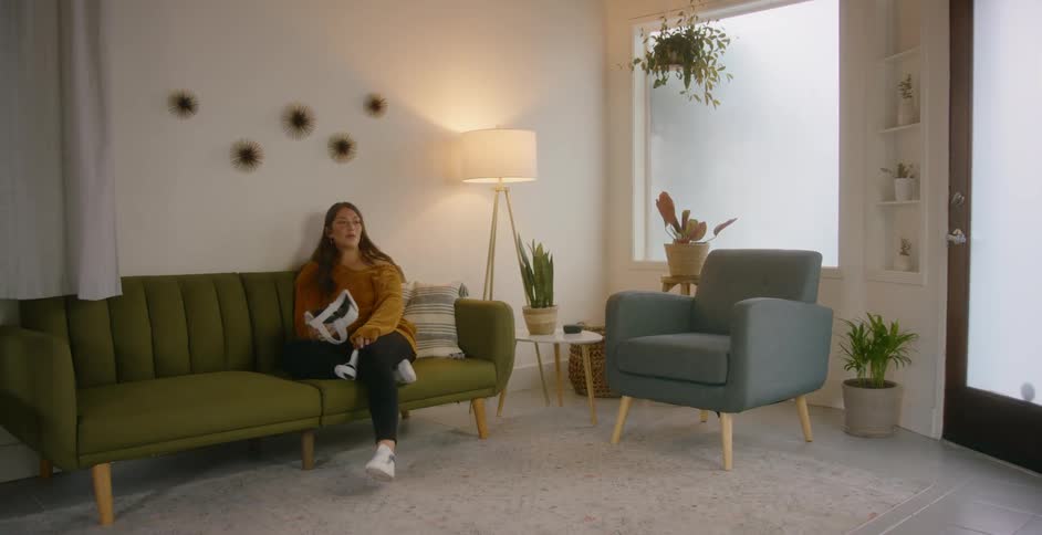 Woman with a brown sweater black leggings and white shoes takes off virtual reality headset and sits down on green couch in the living room at home