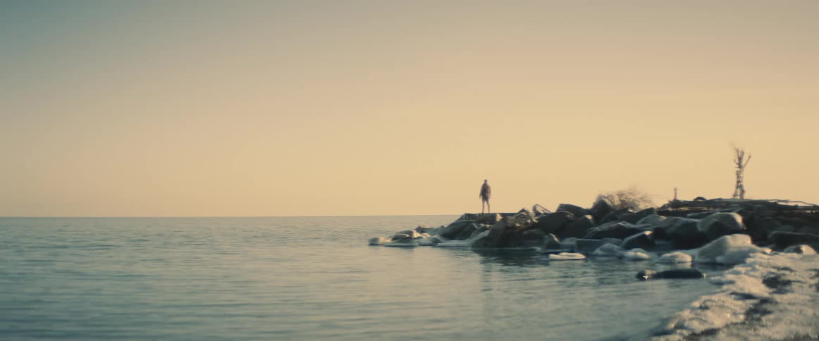 A single person stands at the edge of a shoreline as relatively calm water flows past